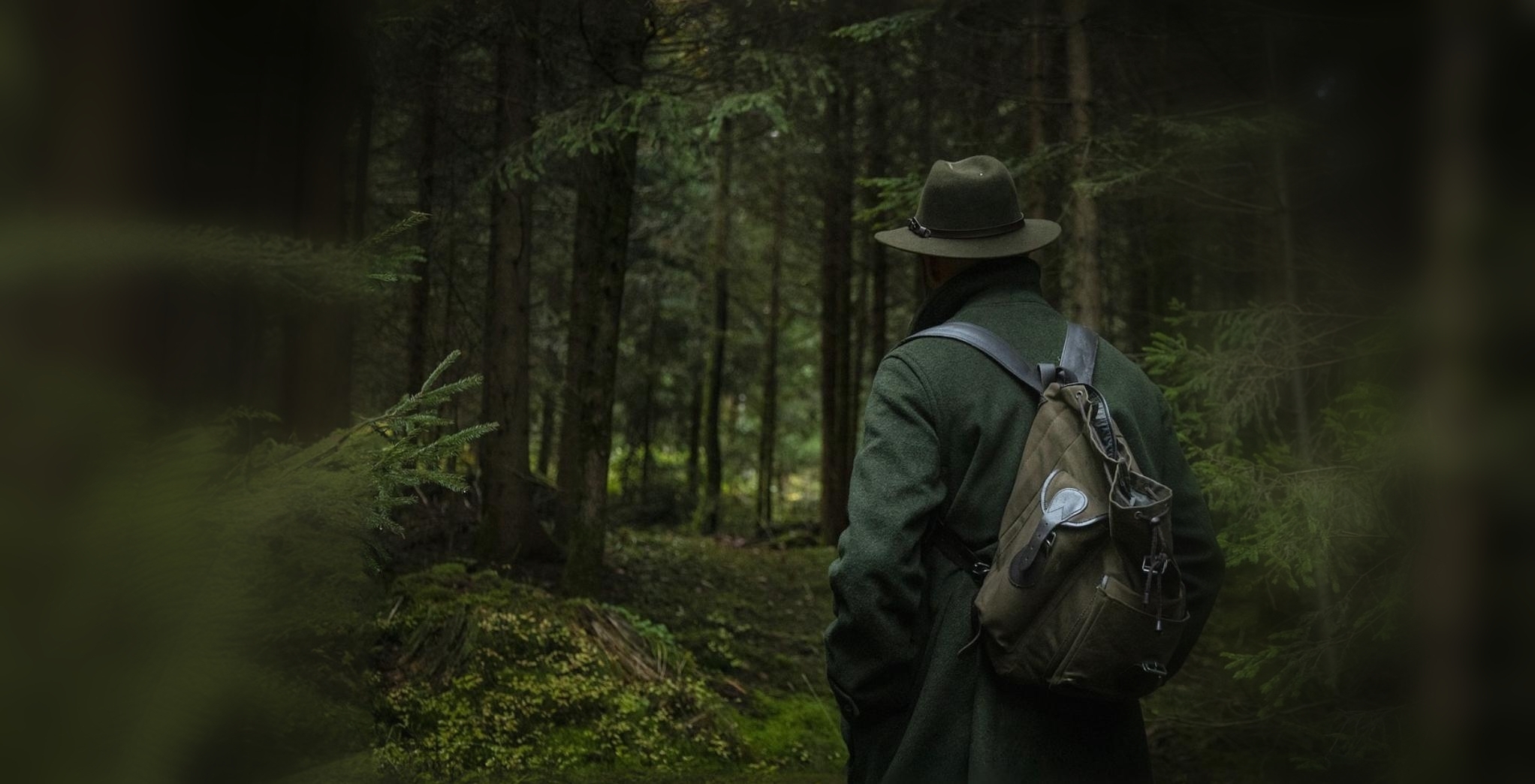 Ein Jäger schaut in den Wald hinein, man sieht ihn von hinten mit Hut und Rucksack, umrandet von Bäumen und Ästen.